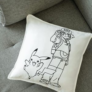 May include: White square pillow featuring a black line drawing of a cartoon character and a Pikachu. The cartoon character is wearing a hat and backpack. Pikachu is jumping with a lightning bolt tail.
