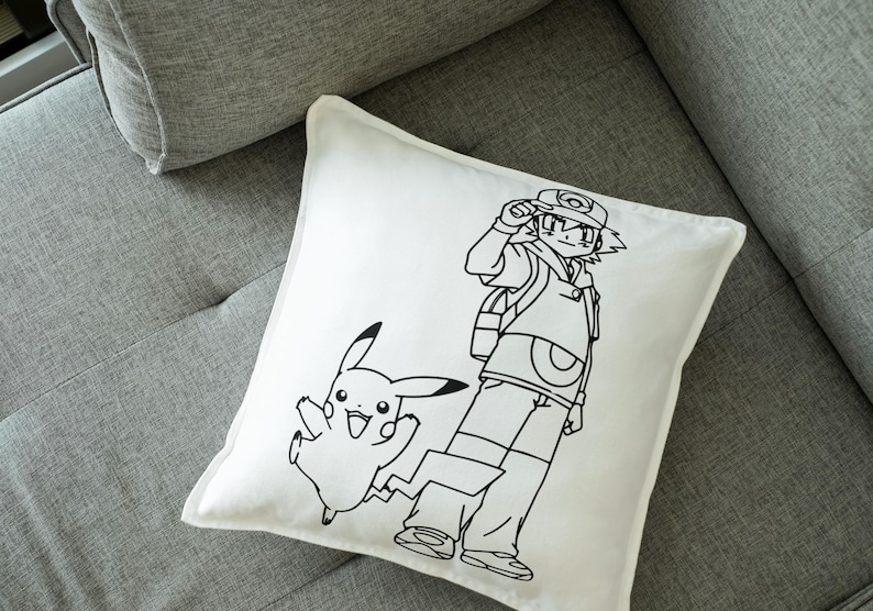 May include: White square pillow featuring a black line drawing of a cartoon character and a Pikachu. The cartoon character is wearing a hat and backpack. Pikachu is jumping with a lightning bolt tail.