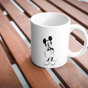 May include: White ceramic mug featuring a black and white illustration of Mickey Mouse holding a heart. The mug has a curved handle and is set on a wooden surface. A classic Disney-themed item.