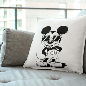 May include: White square pillow featuring a black and white illustration of Mickey Mouse wearing sunglasses. The cartoon character is in a standing pose with his hands on his hips. The pillow is on a gray couch.