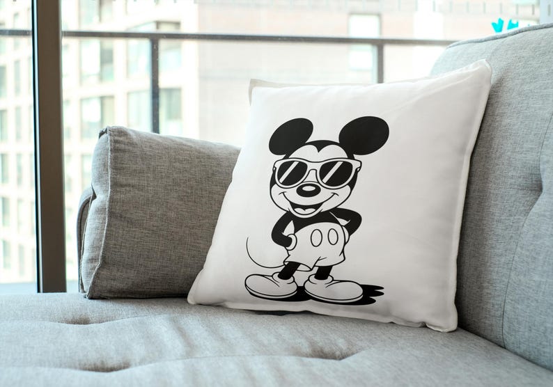 May include: White square pillow featuring a black and white illustration of Mickey Mouse wearing sunglasses. The cartoon character is in a standing pose with his hands on his hips. The pillow is on a gray couch.
