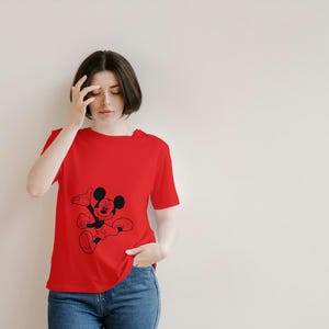 May include: A red t-shirt featuring a black graphic of Mickey Mouse. The shirt is short-sleeved and has a crew neck. The person is wearing blue jeans.