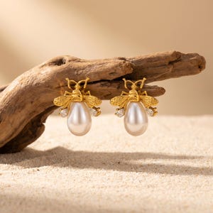 May include: A pair of gold-colored bee-shaped earrings with pearl-shaped drops. The earrings have small, clear gemstones and are displayed on a piece of driftwood against a sandy background.