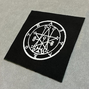 May include: Black square patch featuring a white occult symbol within a circle. The symbol includes a star, lines, and letters. The patch is likely made of fabric and is displayed on a gray surface.