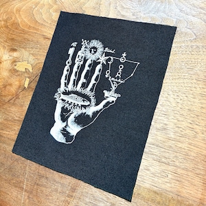 May include: Black rectangular patch featuring a white illustration of a hand holding various symbols, including a sun, keys, and a fish. The design includes the text "AST" and other symbols. The patch is likely for clothing or accessories.