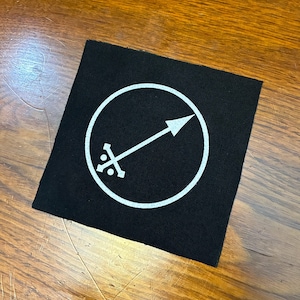 Op de afbeelding: Vierkante zwarte patch met een wit cirkelvormig ontwerp. Het ontwerp toont een witte pijl die diagonaal over een witte cirkel wijst. Onder de pijl bevindt zich een wit kruisachtig symbool. De patch ligt op een houten oppervlak.