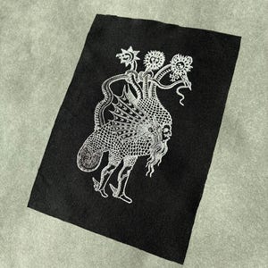 Op de afbeelding: Zwart rechthoekig embleem met een witte illustratie van een mythisch wezen. Het wezen heeft meerdere hoofden, een visachtig lichaam en menselijke benen. Het ontwerp is gedetailleerd met ingewikkelde patronen en lijnen.