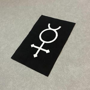 May include: A black rectangular patch with a white alchemical symbol. The symbol features a circle with a curved top and a cross-like shape at the bottom. The patch is made of a textured fabric and is set against a gray background.