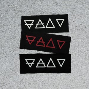 Op de afbeelding: Drie zwarte rechthoekige patches met witte en rode geometrische symbolen. De bovenste en onderste patches hebben witte symbolen, de middelste rode. De symbolen zijn gestileerde representaties van de vier elementen.