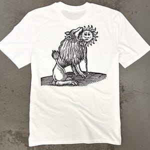 Könnte beinhalten: Weißes T-Shirt mit einer Schwarz-Weiß-Illustration eines löwenartigen Wesens, das eine Sonne mit Gesicht beißt. Die Grafik befindet sich auf der Rückseite. Das T-Shirt hat kurze Ärmel und einen Rundhalsausschnitt.