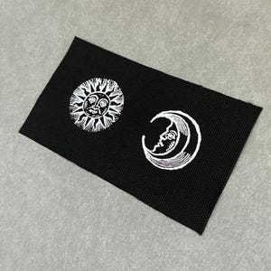 Op de afbeelding: Een rechthoekige zwarte stoffen patch met een wit geborduurd ontwerp van een zon met een gezicht aan de linkerkant en een maansikkel met een gezicht aan de rechterkant. De patch heeft een geweven textuur en strakke randen met een contrastrijke hemelse afbeelding.