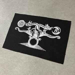 Może przedstawiać: Czarna naszywka z białym rysunkiem dwugłowego smoka, słońca i księżyca. Smok znajduje się nad pierścieniem i trójkątem. Projekt jest szczegółowy i skomplikowany.