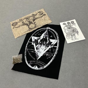 May include: A black fabric patch featuring a white occult illustration of a figure within an oval frame with the text Qvod Superior Macroprosopus and Qvod Inferius, a rectangular paper print of a dragon, a white paper print of a creature, and a small bag.