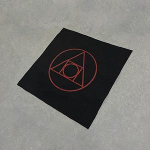 May include: Black square patch with a red geometric design. The design features a circle with a triangle inside, and another triangle and circle inside that. The patch is made of a textured fabric.
