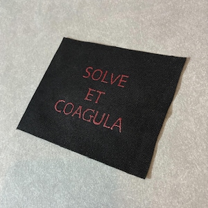 May include: A square, black fabric patch with the words "SOLVE ET COAGULA" embroidered in red. The patch is centered in the image and appears to be made of a woven material.