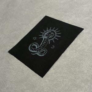 May include: Black rectangular patch featuring a light blue illustration of a snake coiled around a sun with a lion's head, a star, and a crescent moon. The patch is made of a textured fabric.