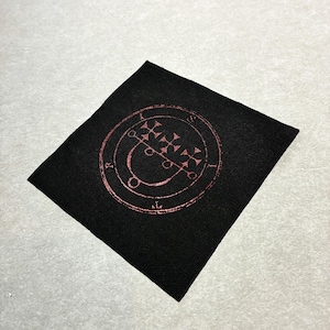 Op de afbeelding: Een vierkante zwarte stoffen patch met een cirkelvormig ontwerp in een metallic koperkleur. De centrale afbeelding bestaat uit een reeks kruissymbolen en gebogen lijnen binnen een ring met de letters R, I en S.