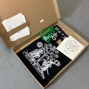 May include: An open cardboard box containing occult items. Inside are cards, a black cloth with a white illustration, a small green bag, and other black and white items. The box is on a grey surface.