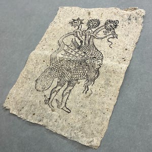 May include: A rectangular piece of textured, off-white handmade paper featuring a black ink illustration of a mythical creature. The creature has a human-like face, scaled body, wings, and three distinct heads resembling a bird, sun, and moon.