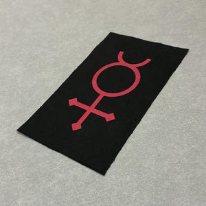 May include: A rectangular black fabric patch featuring a centered, bold red graphic symbol. The design consists of a circle with a crescent arch on top and a cross with pointed ends extending from the bottom, creating a distinct geometric emblem.