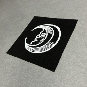 Può includere: Una toppa quadrata in tessuto nero con una grafica serigrafata bianca raffigurante una luna crescente con un volto umano. Il design mostra linee dettagliate e ombreggiature all'interno della forma della luna su un fondo in tessuto nero.