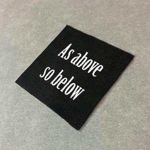 Può includere: Un'etichetta rettangolare nera con la scritta bianca "As above so below". Il testo è scritto in corsivo. L'etichetta è posta su una superficie grigia.