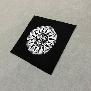 Pode incluir: Um emblema de tecido quadrado preto com um gráfico serigrafado em branco de um sol com um rosto humano. O sol tem raios estilizados que se estendem para fora num padrão circular, centrado no material têxtil preto com textura visível.