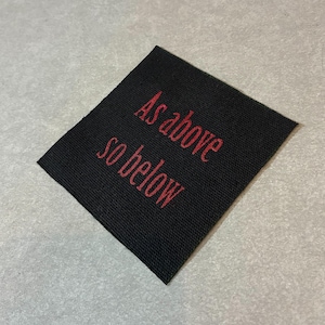 May include: Black woven patch with the red text "As above so below". The patch is rectangular and slightly angled, with the text arranged in two lines. The background is a neutral gray.
