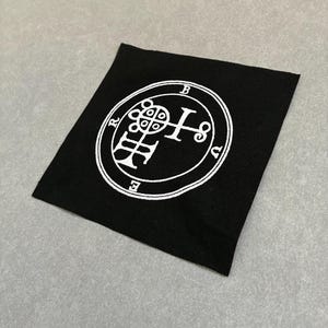 May include: A black square patch with a white circular emblem. The emblem features a complex design with letters and symbols. The patch is made of a textured fabric and is set against a gray background.