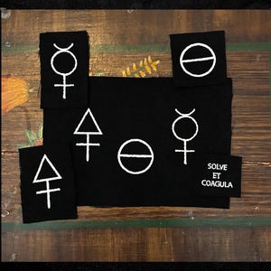 May include: A collection of black fabric patches with white alchemical symbols and the phrase "SOLVE ET COAGULA". The patches feature geometric shapes and symbols, suitable for sewing or crafting projects.
