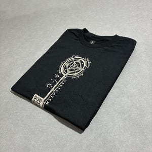 Può includere: Una t-shirt nera a maniche corte con una grande grafica color bianco sporco raffigurante una chiave. La testa della chiave contiene un disegno geometrico con testo in latino, incluse le parole Veritatem, Intellectus e Judicer, mentre il gambo mostra simboli matematici.