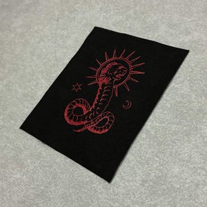 May include: Black rectangular patch with a red embroidered design. The design features a snake with a sunburst, stars, and a crescent moon. The patch is likely for clothing or accessories.