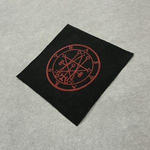 Pode incluir: Tecido quadrado preto com um design circular vermelho com uma estrela e texto. O design está centrado no tecido. O tecido está sobre uma superfície cinza. O design parece ser um símbolo.