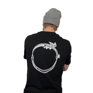 Può includere: T-shirt nero con un disegno bianco di Ouroboros sul retro. L'Ouroboros è un simbolo circolare che raffigura un serpente che si mangia la coda. La persona nell'immagine indossa un berretto grigio.