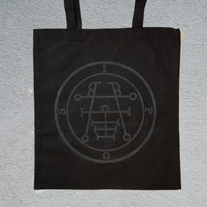 Puede incluir: Bolso tote negro con dos asas. El bolso presenta un diseño circular gris con las letras "I", "B", "P" y "S" alrededor del perímetro. El diseño también incluye un símbolo central y otras formas geométricas.