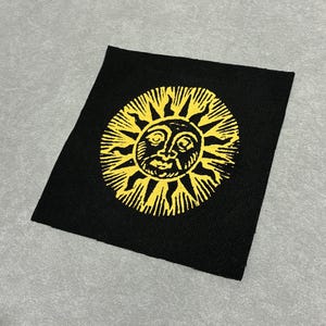 May include: A square black fabric patch featuring a screen-printed yellow sun design. The sun has a central face with detailed facial features and radiating pointed rays extending outward in a circular pattern across the center of the black textile.