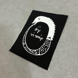 May include: A rectangular black fabric patch featuring a white screen-printed graphic of an Ouroboros, a serpent eating its own tail. Inside the circular shape, the Greek text "ἓν τὸ πᾶν" is printed in white, set against the black background.