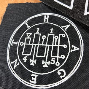 Op de afbeelding: Zwarte stoffen patch met een wit cirkelvormig ontwerp met symbolen, cijfers en letters. Een wit lint met driehoekige vormen en letters staat op de achtergrond. De patch heeft een gedetailleerd, ingewikkeld ontwerp.