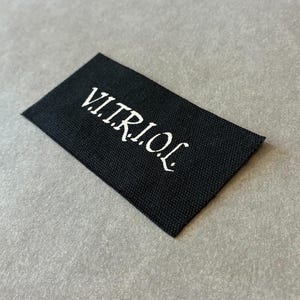 Puede incluir: Una etiqueta rectangular negra con la palabra "V.I.T.R.I.O.L" en blanco, con una tipografía estilizada. La etiqueta tiene una textura y se encuentra sobre un fondo gris neutro. El texto es el foco principal.