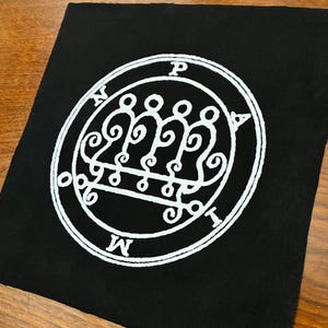 Può includere: Toppa quadrata nera con un disegno circolare bianco. Il disegno presenta simboli intricati e le lettere P, N, M e I. L'immagine include figure stilizzate e volute ornate, creando un'immagine dettagliata e simbolica.