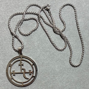 Könnte beinhalten: Eine silberfarbene Metallhalskette mit einem kreisförmigen Anhänger im Scherenschnitt-Design. Der Anhänger zeigt ein zentrales kreuzartiges Symbol mit geschwungenen Elementen in einem Doppelringrahmen, befestigt an einer runden Boxkette.