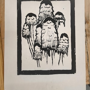 May include: A monochrome linocut print depicting a group of stylized mushrooms. The image is framed by a thick black border, with detailed rendering of the mushroom caps and stems. The print is set against a white backdrop.