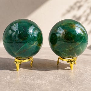 Op de afbeelding: Twee gepolijste donkergroene stenen bollen met zichtbare natuurlijke aders en insluitingen. Elke bol wordt gepresenteerd op een kleine, driepotige goudkleurige metalen standaard. De oppervlakken van de bollen zijn glad en reflecterend.