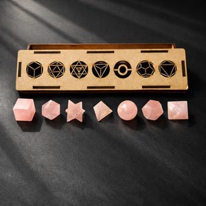 Puede incluir: Una caja de madera con recortes geométricos y un conjunto de formas de cristal de cuarzo rosa. Las formas incluyen un cubo, un hexágono, una estrella, una pirámide, una esfera y otros poliedros. La caja está abierta, revelando los cristales.