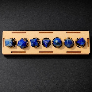 May include: A wooden display tray featuring six blue lapis lazuli gemstones in different geometric forms. Each stone is set within a gold-coloured, circular frame. The rectangular tray has cut-out sections and rests on a dark surface.