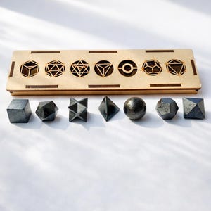 Puede incluir: Un conjunto de siete formas geométricas metálicas de color gris oscuro, que incluyen un cubo, una esfera y una pirámide. Se exhiben sobre una superficie blanca con una caja de madera con recortes geométricos. Las formas varían en tamaño y complejidad.