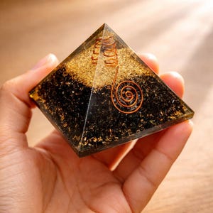 Op de afbeelding: Een heldere hars piramide met een koperen spiraal en gouden vlokken. De piramide heeft een zwarte basis en wordt in een hand gehouden. De koperen spiraal heeft de vorm van een spiraal.