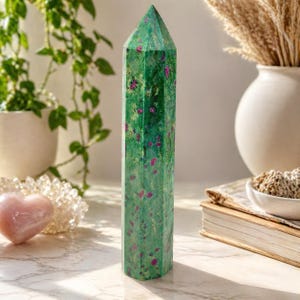 May include: A tall, faceted green crystal tower with red inclusions. The gemstone is displayed on a white surface, with a blurred background featuring plants, a heart-shaped stone, and a vase with dried flowers. The crystal is a decorative item.