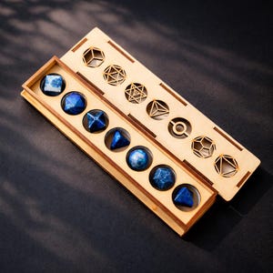 Puede incluir: Un juego de siete dados de piedras preciosas azules en una caja de madera. Los dados tienen varias formas geométricas. La caja tiene una tapa corredera con recortes geométricos. Los dados se muestran sobre una superficie oscura.