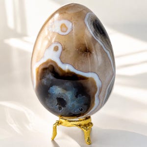 Può includere: Un uovo decorativo in pietra lucida. L'uovo presenta fasce marroni, bianche e nere ed è esposto su un piccolo supporto dorato ornato. Un oggetto di arredamento per la casa unico.